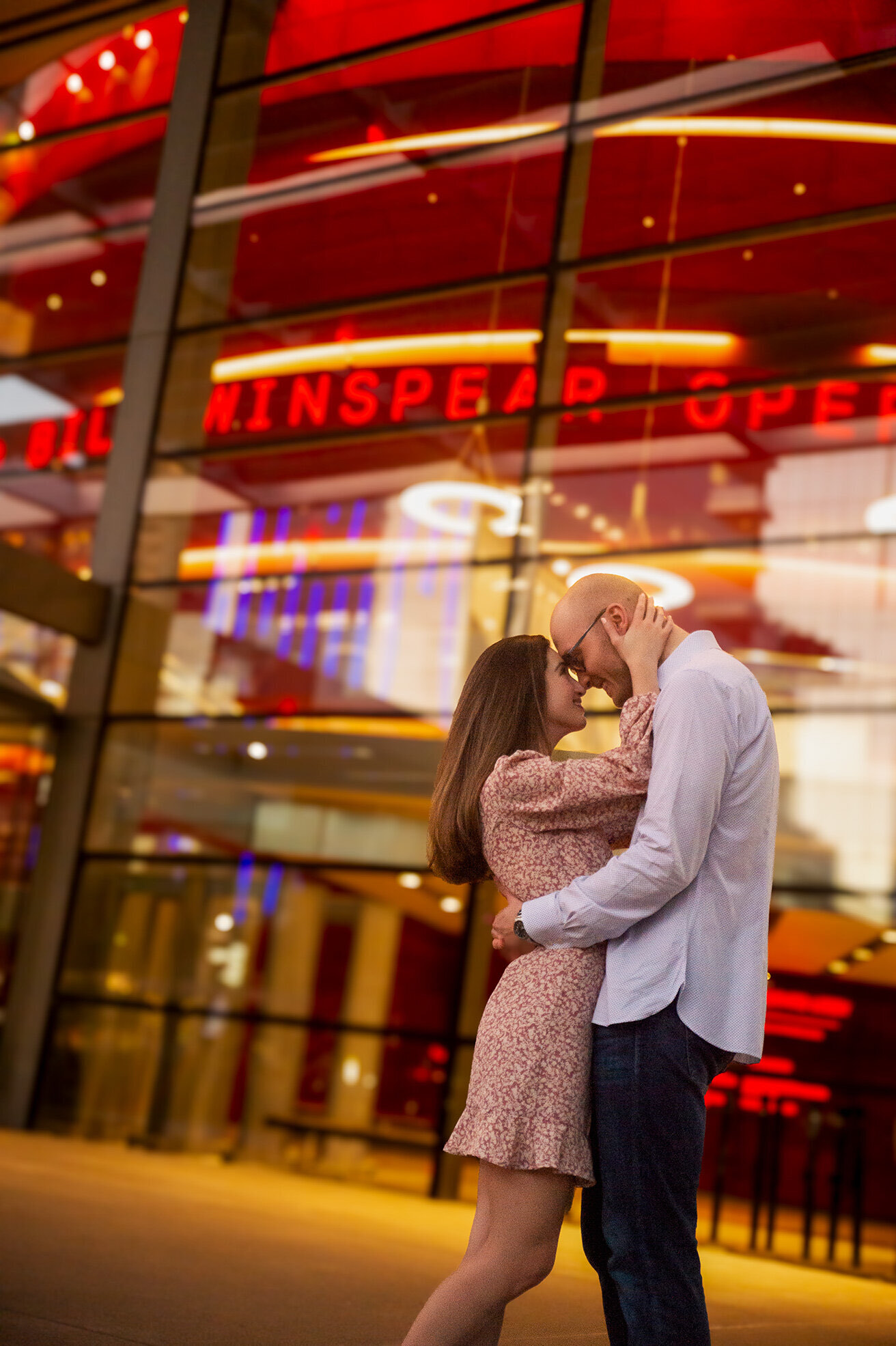 A couple embracing in front of the glowing glass facade of the Winspear Opera House in the Dallas Arts District