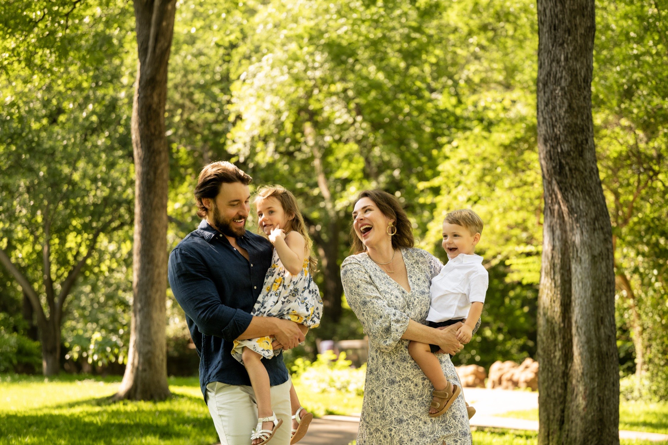 Parents laughing with their two young children under green trees at a Dallas park
