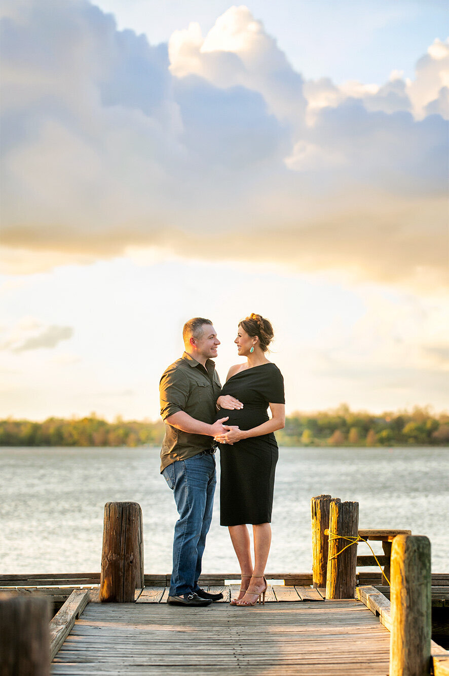 An expecting couple standing together on a wooden dock at White Rock Lake beneath a huge sunset sky